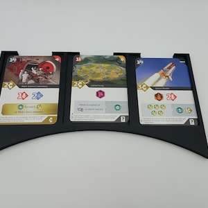 May include: A black, curved card holder with three game cards. The cards feature illustrations of a Mars science laboratory, a landscape, and a space shuttle. The cards have various symbols and text, including numbers and phrases like "for each" and "Have a signal or".