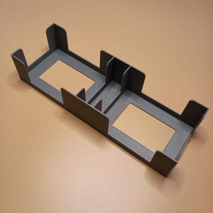May include: A gray 3D-printed organizer with two rectangular openings and several vertical dividers. The organizer has a rectangular shape with raised edges and corner supports. The object is designed to hold small items.