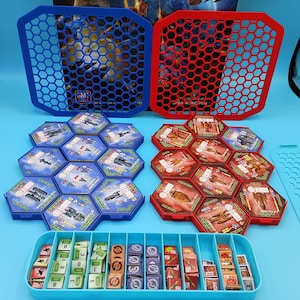 May include: A board game set with blue and red components. The set includes hexagonal tiles, trays, and storage containers. The blue components have the text "GM" and the red components have the text "JIM KROAN". The game pieces are organized in a blue tray.