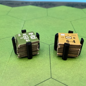 May include: Two stacks of game pieces, each held by a black plastic holder, sit on a green hexagonal board. The game pieces are small, rectangular, and have printed text and symbols. One stack is green, the other is tan.