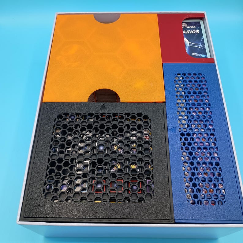 K&ouml;nnte beinhalten: Eine wei&szlig;e Box mit schwarzem, orangefarbenem, rotem und blauem Innenraum. Der Innenraum hat ein Wabenmuster und eine Karte mit dem Text "The Things ARIOS". Die Box ist f&uuml;r ein Brettspiel konzipiert.