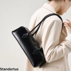 May include: A black rectangular handbag with a top handle and a shoulder strap. The bag is made of a shiny material and has a zipper closure. The word "Standartus" is printed at the bottom of the image.