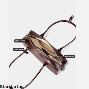 May include: A brown leather handbag with an open zipper revealing the interior. The bag has two handles and features a zipper pocket, a phone pocket, and a main pocket. The word "Standartus" is printed at the bottom.