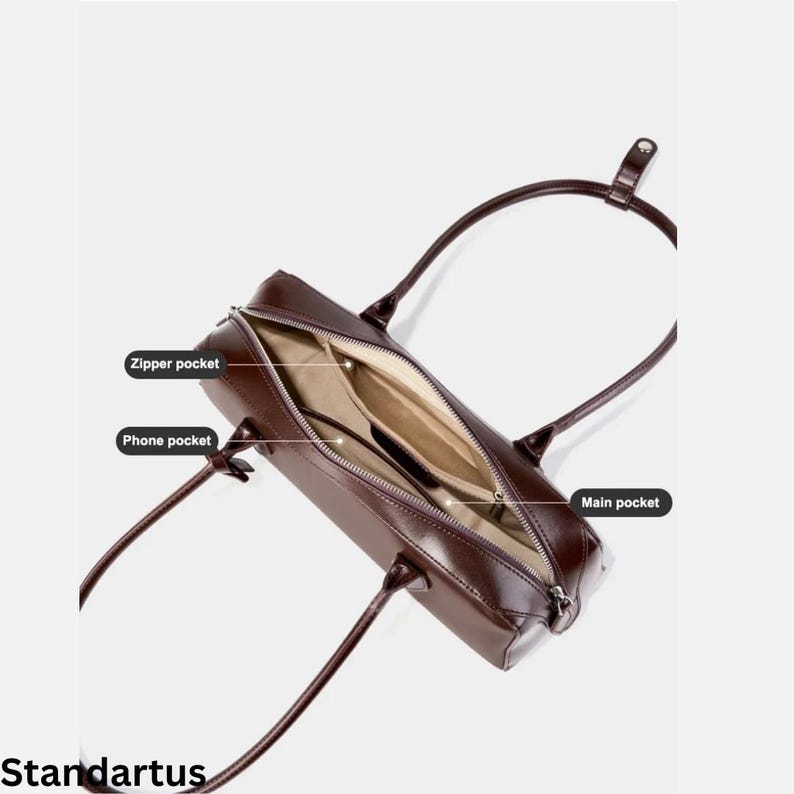 May include: A brown leather handbag with an open zipper revealing the interior. The bag has two handles and features a zipper pocket, a phone pocket, and a main pocket. The word "Standartus" is printed at the bottom.