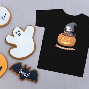 Może przedstawiać: Czarny t-shirt z halloweenowym motywem przedstawiającym postać w kapturze czytającą w dyni. Ciasteczka na Halloween w kształcie ducha, nietoperza, dyni i okrągłe ciasteczka z napisem "Boo!".
