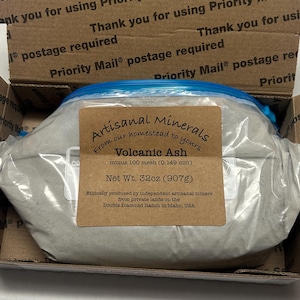 May include: A package of volcanic ash, labeled "Artisanal Minerals," inside a cardboard box. The label indicates a net weight of 32oz (907g). The box is marked with "Priority Mail" postage information.