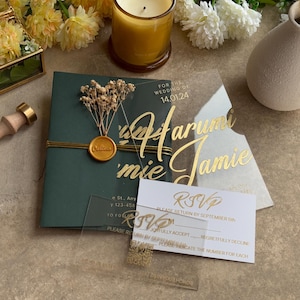 Op de afbeelding: Een elegante trouwuitnodigingssuite met een donkergroene uitnodigingskaart met gouden accenten, een gouden zegel en een transparante acryl overlay met de namen "Harumi & Jamie". Inclusief RSVP-kaarten.