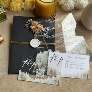 May include: A stylish wedding invitation set. It includes a black envelope with gold detailing, clear acrylic invitations, and RSVP cards. The design features a forest scene and calligraphy. A wax seal and dried flowers add a decorative touch.