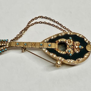 Pode incluir: Broche de bandolim vintage em tom dourado com detalhes em turquesa e cristal transparente. O broche apresenta um fundo de esmalte azul escuro e uma corrente delicada. Uma peça de joalheria clássica com tema de instrumentos musicais.