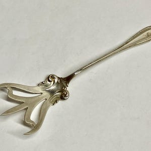 Puede incluir: Un tenedor de servir plateado con un mango decorativo y un diseño calado único en las púas. El tenedor presenta un patrón adornado y enrollado y un monograma en el mango. El utensilio está diseñado para servir alimentos.
