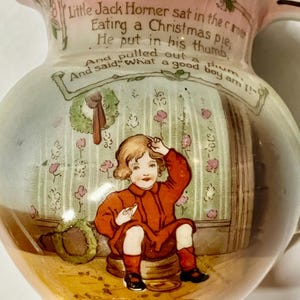 Könnte beinhalten: Vintage Keramikkrug mit rosa Rand und detaillierter Illustration. Die Illustration zeigt ein Kind in roter Kleidung mit Text aus einem Kinderreim. Der Krug hat einen Henkel und einen Ausguss und dient wahrscheinlich dekorativen Zwecken.