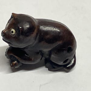 Puede incluir: Figura de madera tallada de color marrón oscuro que representa un gato en reposo. El gato tiene orejas pequeñas y redondas, un hocico corto y ojos visibles. La figura está detallada con un acabado liso y pulido.