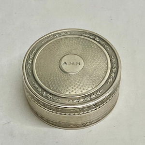 May include: A silver-toned, round trinket box with a detailed floral and leaf design around the rim. The lid features a textured pattern and the initials "A.H.H." in a central circle. The box is likely made of metal.