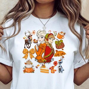 May include: A white t-shirt featuring a colorful graphic print of two animated characters, a reindeer, a snowman, and various autumn-themed elements including pumpkins, a bow, a slice of cake, a mug, and an umbrella, all arranged in a central collage.
