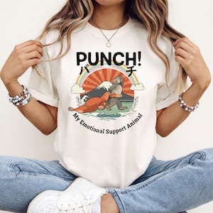 May include: Cream-colored t-shirt featuring a graphic with the word "PUNCH!" above a cartoon illustration of a seal and a creature. The text "My Emotional Support Animal" is below the illustration. The design includes a rainbow, mountain, and sun.