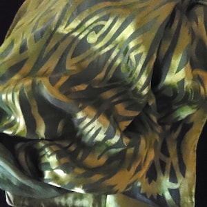 May include: A folded silk scarf with a pattern of gold and olive green abstract shapes. The scarf has a soft, flowing texture and a subtle sheen. The design is reminiscent of organic forms.