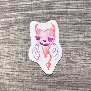 May include: A die-cut sticker featuring a pink heart-shaped character with small red horns, purple closed eyes, a single visible fang, white wings on the sides, and a thin red tail ending in a pointed arrow shape, all outlined in a white border.