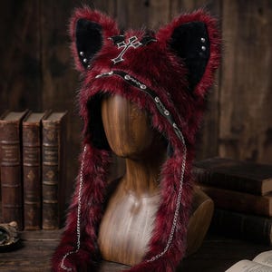 Pode incluir: Um chapéu de pele sintética bordô com detalhes de orelhas pretas e tachas prateadas. O chapéu apresenta um emblema de cruz e um detalhe de corrente. O chapéu é exibido em uma cabeça de manequim de madeira.