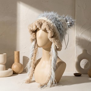 May include: A fluffy, faux fur hat in shades of beige, gray, and white, displayed on a mannequin head. The hat features long, feathery accents and a delicate bow detail. This unique hat is a statement piece.