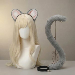 Puede incluir: Una diadema con orejas de gato grises con interiores rosas y blancos, una cola gris a juego y un collar negro con un cierre plateado. Los artículos se exhiben en una cabeza de maniquí con cabello rubio.