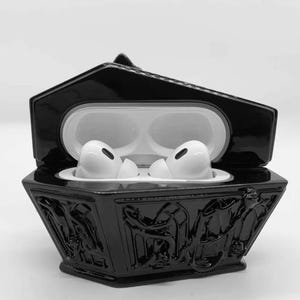 May include: Black coffin-shaped case with a glossy finish, holding white wireless earbuds. The case has intricate relief carvings on the sides and a hinged lid. The earbuds are in a white charging case.