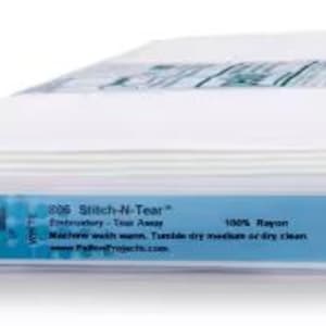 May include: A roll of white Pellon 806 Stitch-N-Tear embroidery fabric. The label is light blue with black text, including "100% Rayon" and "Made in USA". The fabric is tightly rolled and ready for use.
