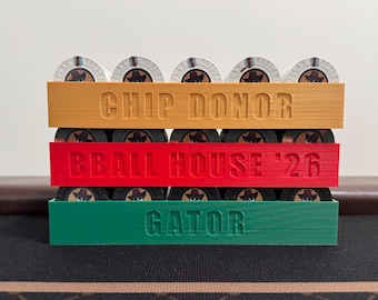 Op de afbeelding: Een drielaagse pokerchiphouder in oranje, rood en groen. Elke laag bevat chips met een cowboyhoedontwerp. De bovenste laag leest "CHIP DONOR", de middelste "BBALL HOUSE '26" en de onderste "GATOR".