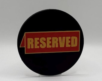 Op de afbeelding: Een zwart, cirkelvormig bord met het woord "RESERVED" in gouden letters op een rode rechthoekige achtergrond. Het bord wordt waarschijnlijk gebruikt om aan te geven dat een ruimte of item niet beschikbaar is.