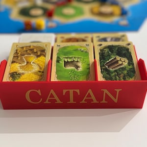 以下が含まれることがあります： 前面に金色の文字でCATANと印刷された赤いプラスチック製のカードホルダー。ホルダーには、小麦、緑の野原にいる羊、森の中の丸太のイラストが描かれたゲームカードが3つの束になって入っており、背景にはゲームボードが見えます。
