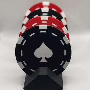 Op de afbeelding: Een rij pokerchips in zwart, rood en wit, gepresenteerd op een zwarte standaard. De voorste chip is zwart met een wit schoppensymbool. De chips hebben witte markeringen rond de randen, wat een klassieke casino-esthetiek creëert.