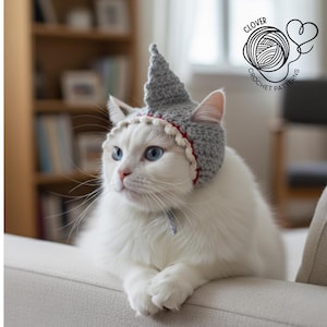 Könnte beinhalten: Eine weiße Katze mit blauen Augen trägt einen gehäkelten grauen Hut mit rotem und weißem Besatz. Die Katze sitzt auf einem hellen Sofa. Der Hut hat eine spitze Spitze. Das Bild enthält auch den Text "CLOVER CROCHET PATTERNS".