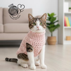 May include: A cat wearing a pale pink, hand-crocheted jumper with a roll neck. The jumper features a textured stitch design. The image includes the text "CLOVER CROCHET PATTERNS" with a yarn ball and heart logo.