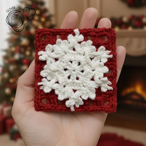 Könnte beinhalten: Ein quadratischer, gehäkelter Weihnachtsschmuck mit einem weißen Schneeflockenmuster auf rotem Grund. Der Schmuck wird in einer Hand gehalten, mit einem verschwommenen Weihnachtsbaum und Kamin im Hintergrund. Das Clover Crochet Pattern Logo ist sichtbar.