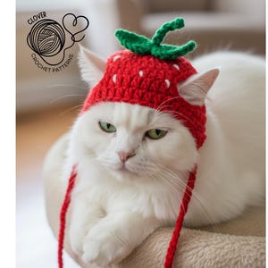 May include: A white cat wearing a red crocheted strawberry hat with green leaves. The hat has white seed details. The cat is sitting on a beige surface. The image includes the text "CLOVER CROCHET PATTERNS".