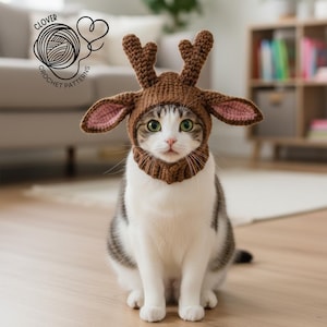 Puede incluir: Un gato con un gorro de reno de ganchillo marrón con cuernos y orejas forradas de rosa. El gato es blanco y gris con ojos verdes, sentado en un suelo de madera. El gorro tiene una pieza para el cuello. Incluye el texto "CLOVER CROCHET PATTERNS".