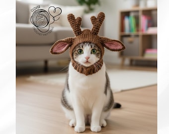 May include: A cat wearing a brown crocheted reindeer hat with antlers and pink-lined ears. The cat is white and gray with green eyes, sitting on a wooden floor. The hat has a neck piece. The image includes the text "CLOVER CROCHET PATTERNS".