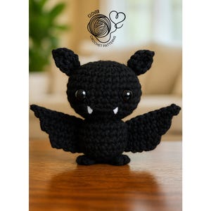May include: A black crochet bat with large, round eyes, small ears, and outstretched wings. The bat has two small white fangs and is sitting on a wooden surface. The image includes the text "Clover Crochet Patterns".