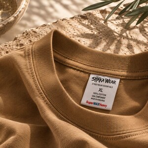 May include: A tan crew neck t-shirt with a ribbed collar. A white interior label reads SHAKA WEAR STREETWEAR ESSENTIALS XL 100% COTTON USA COMPONENT MADE IN HONDURAS SuperMAXHeavy. The fabric has a smooth texture and a solid, earthy brown color.