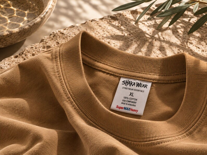 以下が含まれることがあります： リブ編みの襟が付いたベージュのクルーネックTシャツ。白い内側のラベルには、SHAKA WEAR STREETWEAR ESSENTIALS XL 100% COTTON USA COMPONENT MADE IN HONDURAS SuperMAXHeavyと記載されています。生地は滑らかな質感で、落ち着いたアースカラーの茶色です。