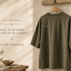 May include: An olive green short-sleeved t-shirt hangs on a wooden hanger. The shirt features the text "PARADISE PALETTE" on the upper left chest. To the left, text reads "A LIVING PEARL", "MADE TO BE WORN. CREATED TO BE CARRIED.", and "INNER FREEDOM".