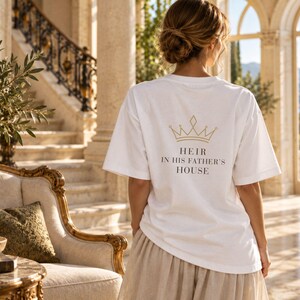 May include: A white short-sleeved t-shirt featuring a gold-colored crown graphic above the black text "HEIR IN HIS FATHER'S HOUSE" printed on the back. The shirt has a crew neckline and a relaxed fit, worn with a light-colored skirt.