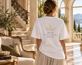 Puede incluir: Una camiseta blanca de manga corta con un gráfico de corona dorada sobre el texto negro "HEIR IN HIS FATHER'S HOUSE" impreso en la espalda. La camiseta tiene cuello redondo y un corte holgado, combinada con una falda de color claro.