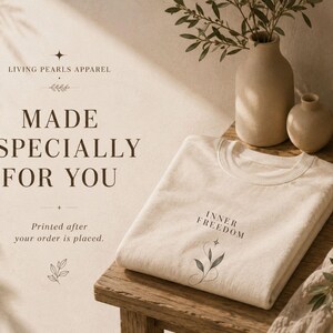 Puede incluir: Una camiseta color crema doblada con el texto negro "INNER FREEDOM" sobre una peque&ntilde;a ilustraci&oacute;n bot&aacute;nica de hojas y una estrella. La camiseta se muestra sobre una superficie de madera junto a dos jarrones de cer&aacute;mica beige y un cuenco con perlas blancas.