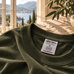 May include: An olive green t-shirt with a ribbed crew neckline. A white interior neck label displays the text SHAKA WEAR, STREETWEAR ESSENTIALS, XL, 100% COTTON, USA COMPONENT, MADE IN HONDURAS, and SuperMAXHeavy in red and blue lettering.