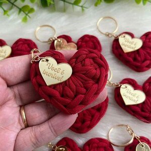 May include: Red heart-shaped keychains with gold-colored key rings and tags. The tags have the text "Voc&ecirc; &eacute; incr&iacute;vel" in gold lettering. The hearts are made of a textured, woven material.