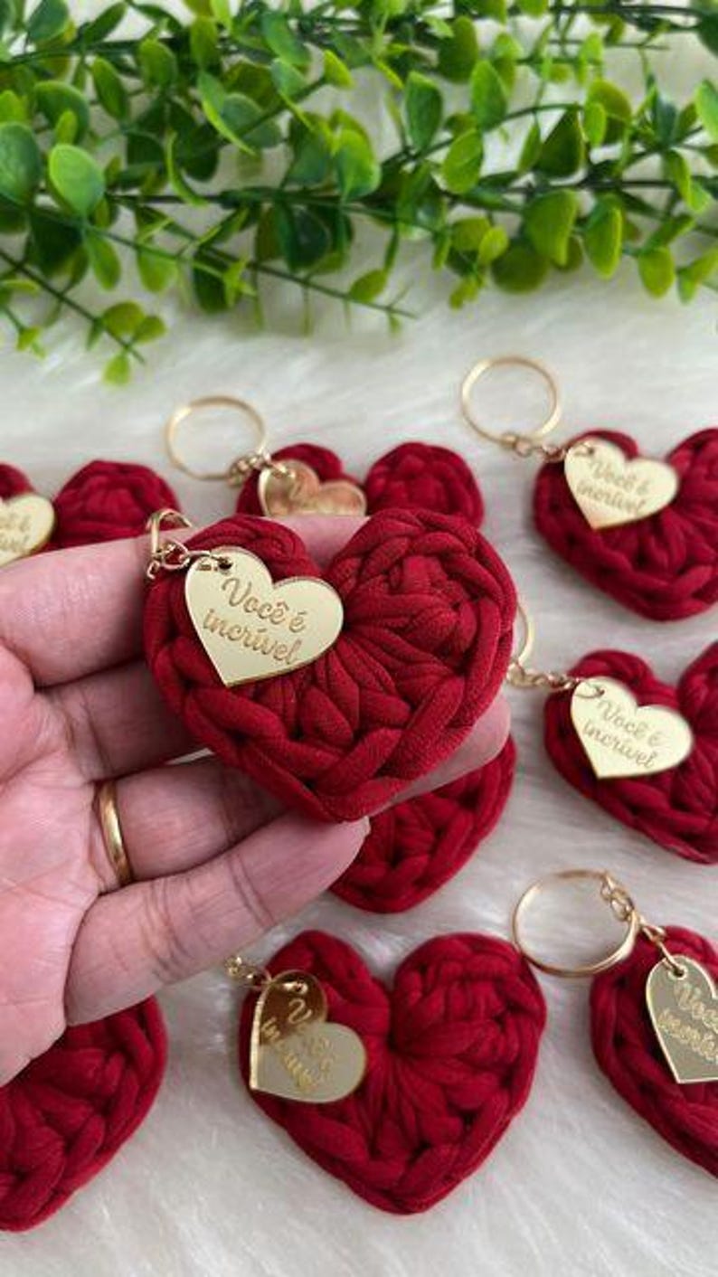 May include: Red heart-shaped keychains with gold-colored key rings and tags. The tags have the text "Voc&ecirc; &eacute; incr&iacute;vel" in gold lettering. The hearts are made of a textured, woven material.