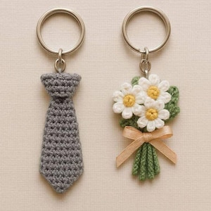Op de afbeelding: Twee sleutelhangers met zilveren ringen. Eén heeft een grijze gehaakte stropdas, de andere een boeket witte madeliefachtige bloemen met gele centra, groene bladeren en een beige lint. Beide zijn handgemaakt.