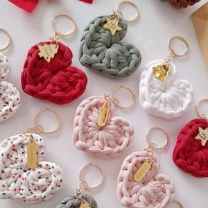 May include: A collection of heart-shaped keychains in various colors, including red, pink, white, and green. Each keychain is crafted from a thick, woven material and features a gold-colored charm with text. The keychains are arranged on a white surface.