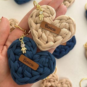 Könnte beinhalten: Herzförmige Schlüsselanhänger in Marineblau und Beige mit goldfarbenen Beschlägen. Jeder Schlüsselanhänger hat ein kleines Lederetikett mit dem Namen "Benício" oder "Florinte" und einen Anhänger. Die Schlüsselanhänger sind aus dickem, gewebtem Material.