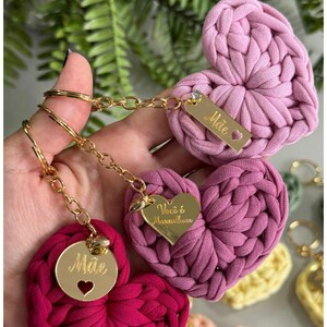 May include: Handmade heart-shaped keychains in shades of pink and red. Each keychain features a gold-colored tag with text and a heart-shaped cutout. The keychains are connected by a gold chain and ring.
